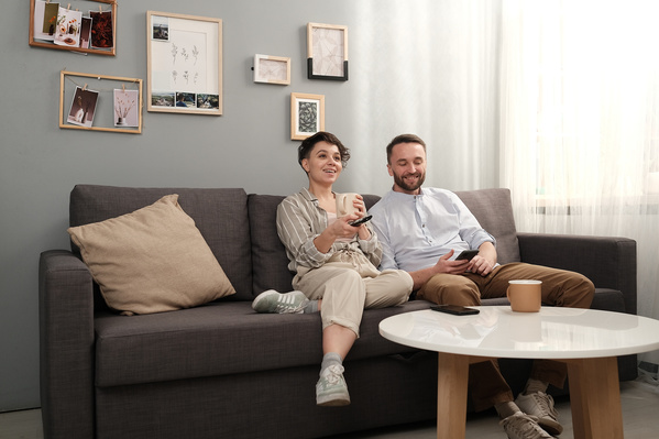 A dark-haired couple in light clothes watching TV in a living room on a gray sofa
