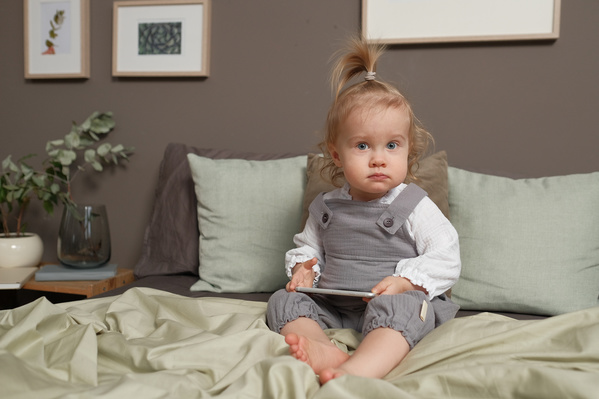 A little blonde girl in a gray jumpsuit with a silver phone sitting on the bed
