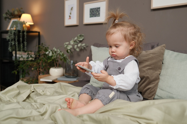 A little blonde girl in a gray jumpsuit playing a silver phone on the bed