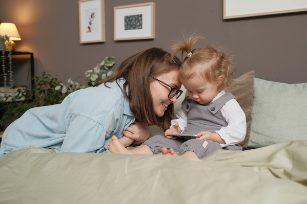 A little blonde girl in a gray jumpsuit watching a cartoon on a silver phone and her mom in glasses and a light shirt on the bed