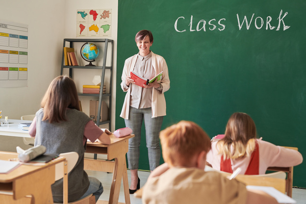 A smiling teacher with short hair dressed in a strict style working with children sitting at desks in a classroom with a large chalkboard