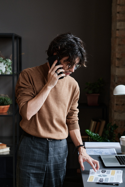 A man with dark curly hair wearing a brown jumper discussing a document on the phone