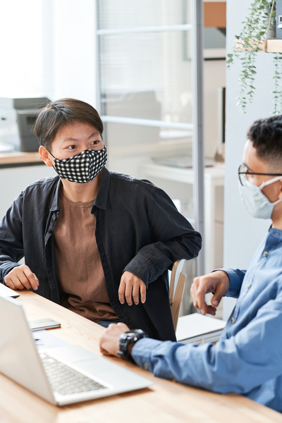 A dark-haired office worker in a reusable mask and a black shirt talking to a colleague