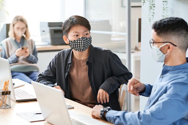A dark-haired man in a reusable mask and a black shirt communicating with a colleague