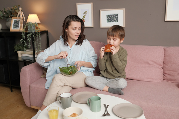 A woman in a light shirt stirring a vegetable salad and her son in pajamas eating a sweet bun while sitting on a pink sofa