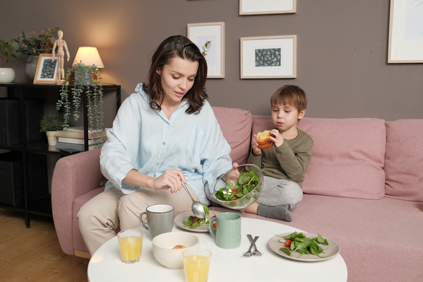 A woman in a light shirt putting vegetable salad on plates and her son in pajamas eating a sweet bun while sitting on a pink sofa