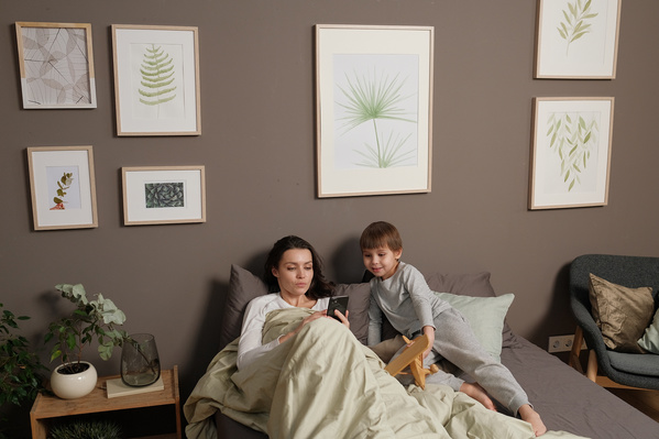 Dark-haired mom in light clothes lying in bed with her little son in pajamas and using the phone
