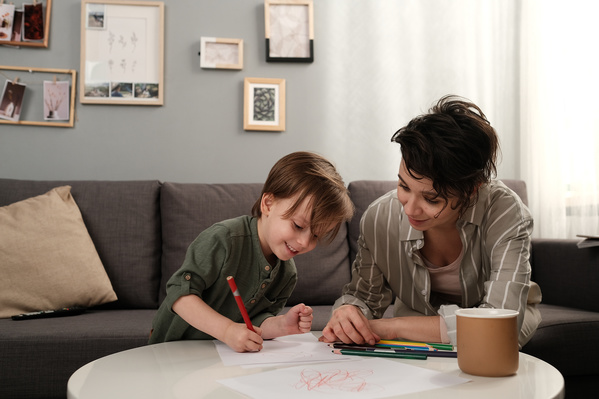 A boy in a green longsleeve drawing pictures with a red pencil and his dark-haired mother watching him and smiling