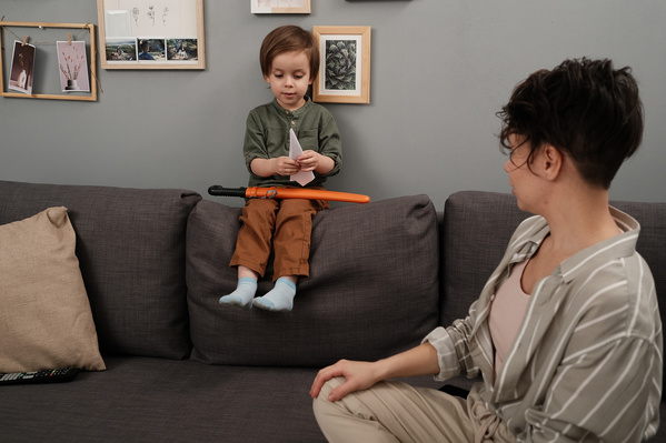 A little boy with a toy making a paper airplane on a gray sofa with his mother