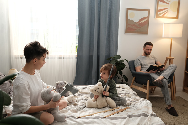 A mother in a light T-shirt playing with toys with her little son in pajamas on the bed and a father reading a book in an armchair