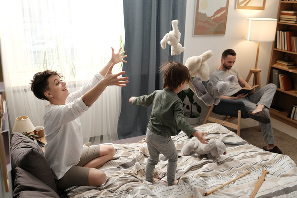 Mom in a light T-shirt tossing toys over her little son in pajamas on the bed