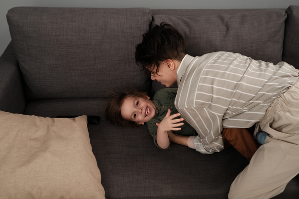 Mom in light clothes tickling her little son in a green longsleeve on a gray sofa