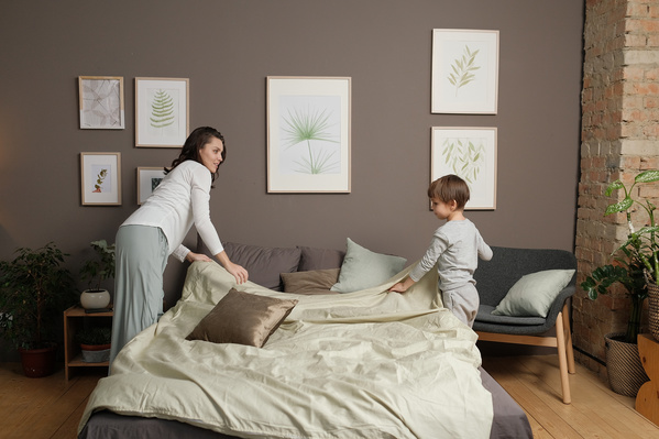 Woman in light clothes and her little son in gray pajamas making the bed