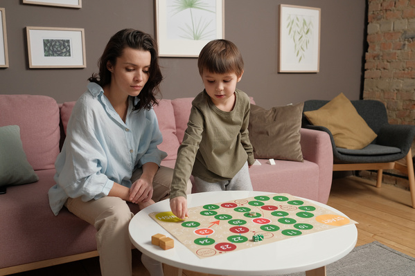 A boy in pajamas playing a board game with his dark-haired mom in light clothes sitting on the couch