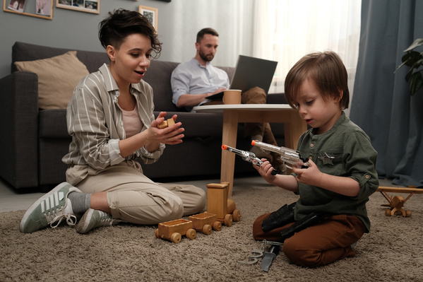 A mom in light clothes playing with her little son with toy guns and a wooden train