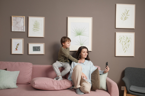 Mom with dark hair in light clothes taking a photo with her little son on a pink sofa