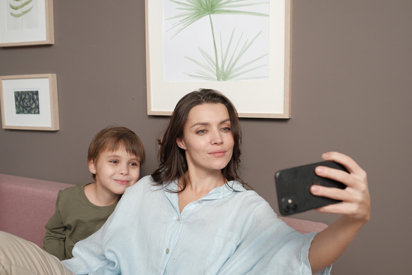 A woman with dark hair in light clothes taking a selfie with her little son on a pink sofa