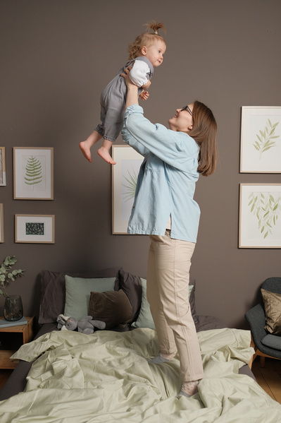 Mom in light clothes lifting her little blonde daughter while standing on an unmade bed