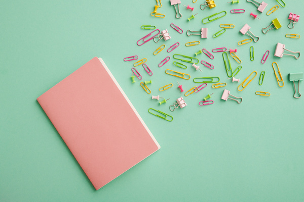Top view of a pink notebook and bright paper clips and pins on a turquoise surface