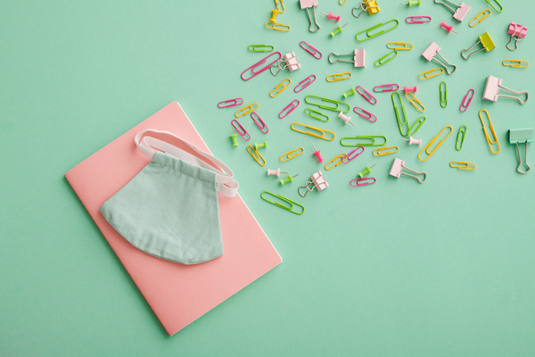 A pink notebook and reusable medical mask next to colored paper clips and pins