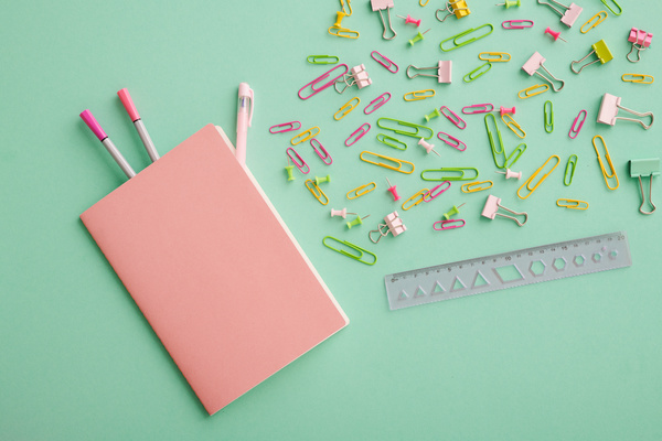 A pink notebook and multicolored felt-tip pens embedded in it next to a ruler and colored paper clips and pins