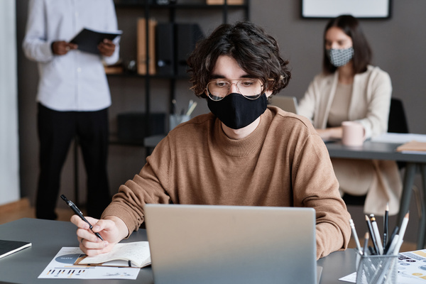 An male employee with dark curly hair wearing a black medical mask working at a desk with a laptop in the office