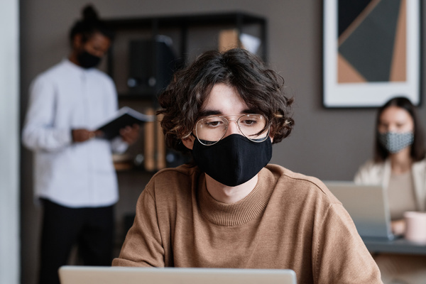 An man with dark curly hair wearing a black face mask working at a desk in the office
