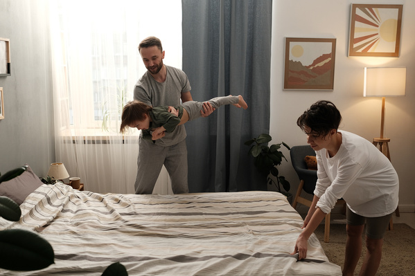 Dark-haired father fooling around with his little son in pajamas and a mom making the bed