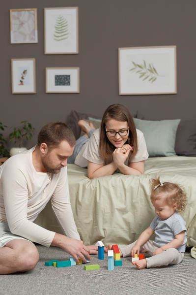 Dad in light pajamas playing with his little blonde daughter with bright wooden blocks and mom watching them from the bed