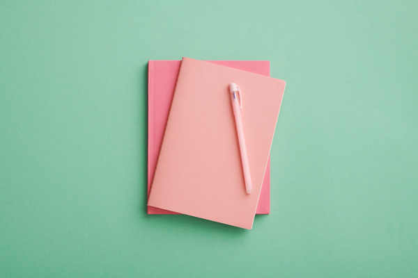 Top view of a pair of pink notebooks and a pen lying on a turquoise surface