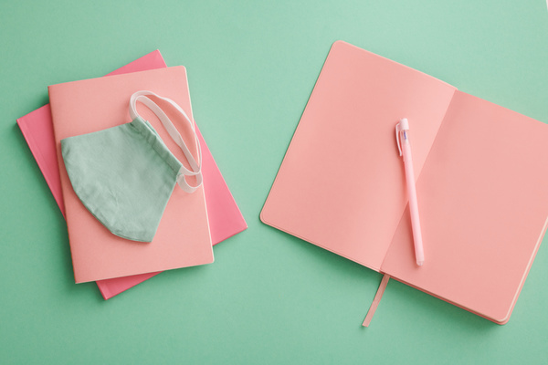 Top view of bright pink notepads with a pen and a reusable medical mask laid out on a turquoise surface