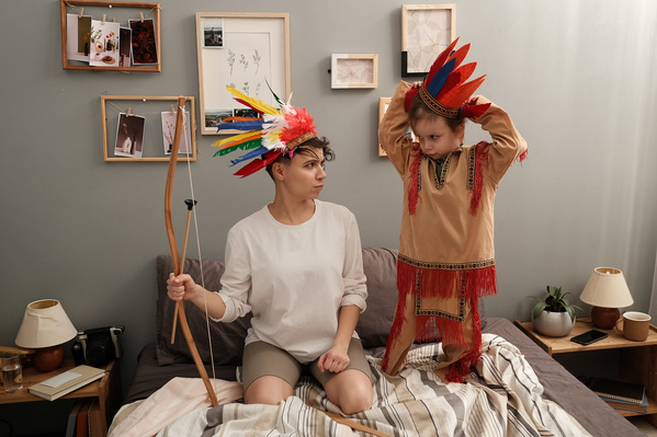 A woman in a white longsleeve and her son in roaches of bright feathers depicting Indians on a bed