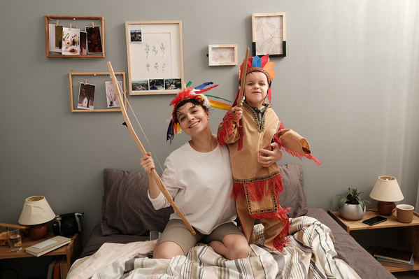 A woman in a white longsleeve and her son in bright-feathered roaches playing Indians on the bed