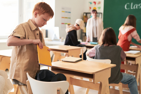 A red-haired schoolboy in a shirt pulling out a yellow notebook for class from a black schoolbag