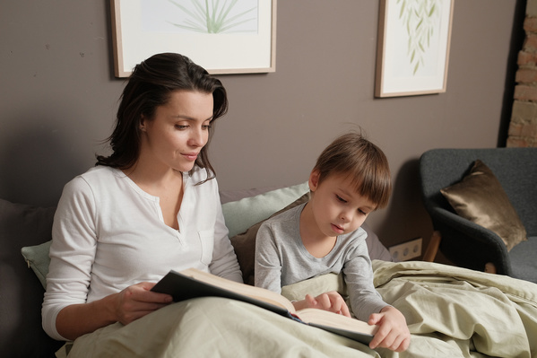 A dark-haired mother in light pajamas reading a book to her little son in bed