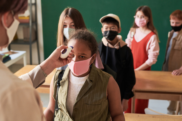 A teacher checking the temperature of a schoolgirl wearing a pink reusable medical mask and entering a class with classmates