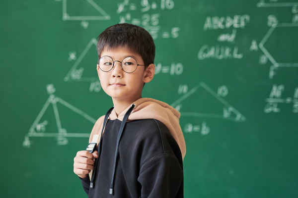 A little schoolboy with dark hair wearing glasses with round lenses and a black hoodie in a classroom