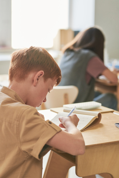 A junior red-haired schoolboy dressed in a beige shirt doing a writing exercise at a desk in the classroom