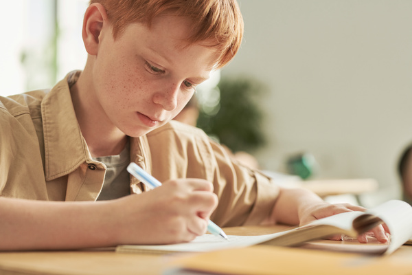 A red-haired middle school student in a beige shirt doing written exercise at his desk
