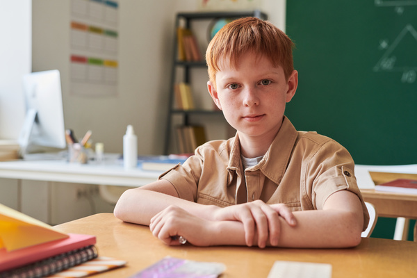 A red-haired schoolboy with freckles dressed in a beige shirt and sitting at a school desk