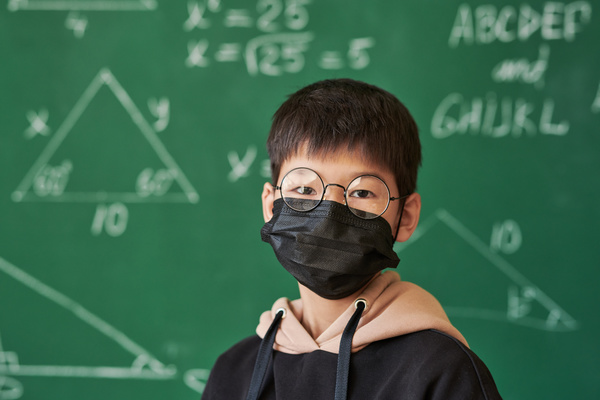 A dark-haired schoolboy in glasses with round lenses and with a black medical mask on his face against a green chalk board