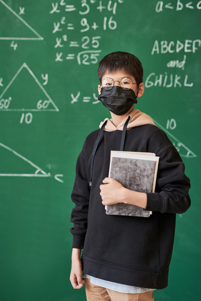 A schoolboy with dark hair wearing glasses with round lenses and with a black medical mask on his face holding a textbook in his hands against a green chalk board