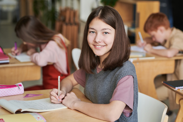 Smiling schoolgirl with brown hair sitting at a wooden desk with a study notebook