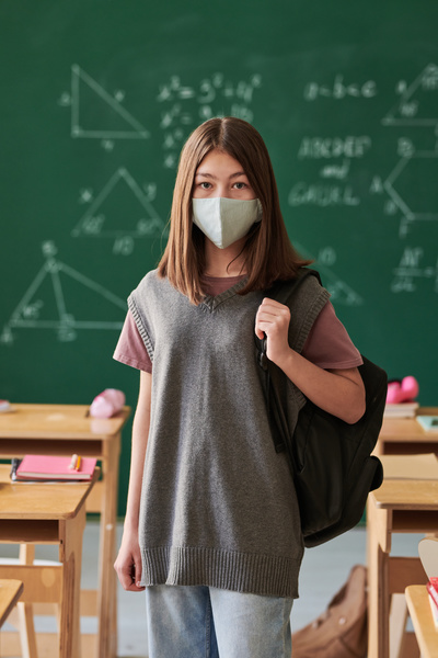 A student with brown hair wearing a reusable light-colored medical mask while in classroom with a chalkboard