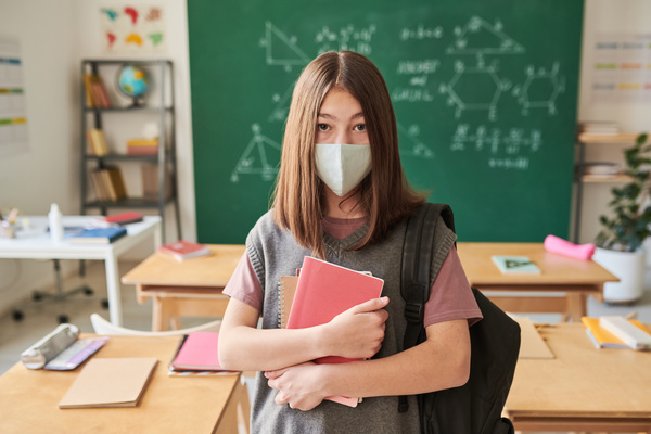 A schoolgirl with brown hair in a reusable light-colored medical mask holding textbooks and notebooks in classroom with a chalkboard