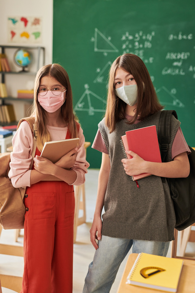 Schoolgirls with brown hair in reusable medical masks of light colors holding textbooks and notebooks