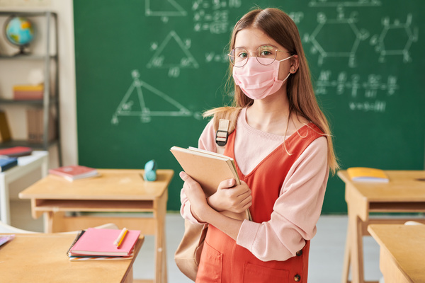 A schoolgirl with long hair in a pink fabric medical mask standing in the classroom with textbooks and notebooks in her hands