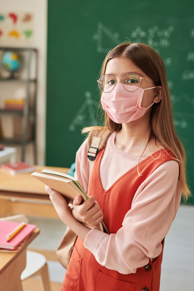 A schoolgirl with long hair in a pink fabric medical mask posing in class with textbooks and notebooks in her hands