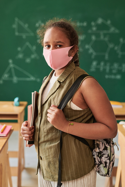 A schoolgirl with tidied hair in a pink fabric face mask with notebooks in her hands and a school bag on her shoulder