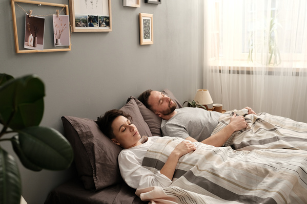 Dark-haired parents in light pajamas sleeping under a white blanket in a bright bedroom